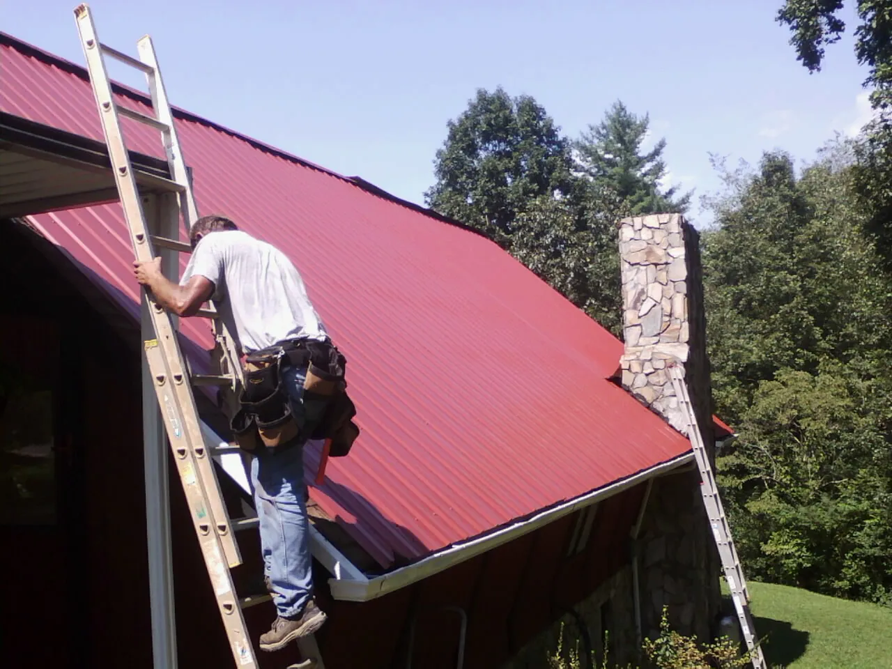 Roofer on a ladder beside a red metal roof and stone chimney — Soffit & Fascia Repair by Roofing Brothers in Government Camp